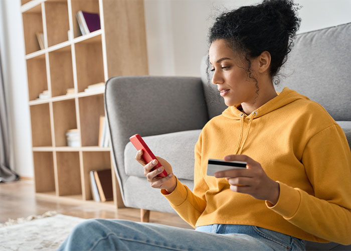 A woman in an orange sweater sits on her living room floor and looks at a smartphone in one hand while holding a credit card in the other.