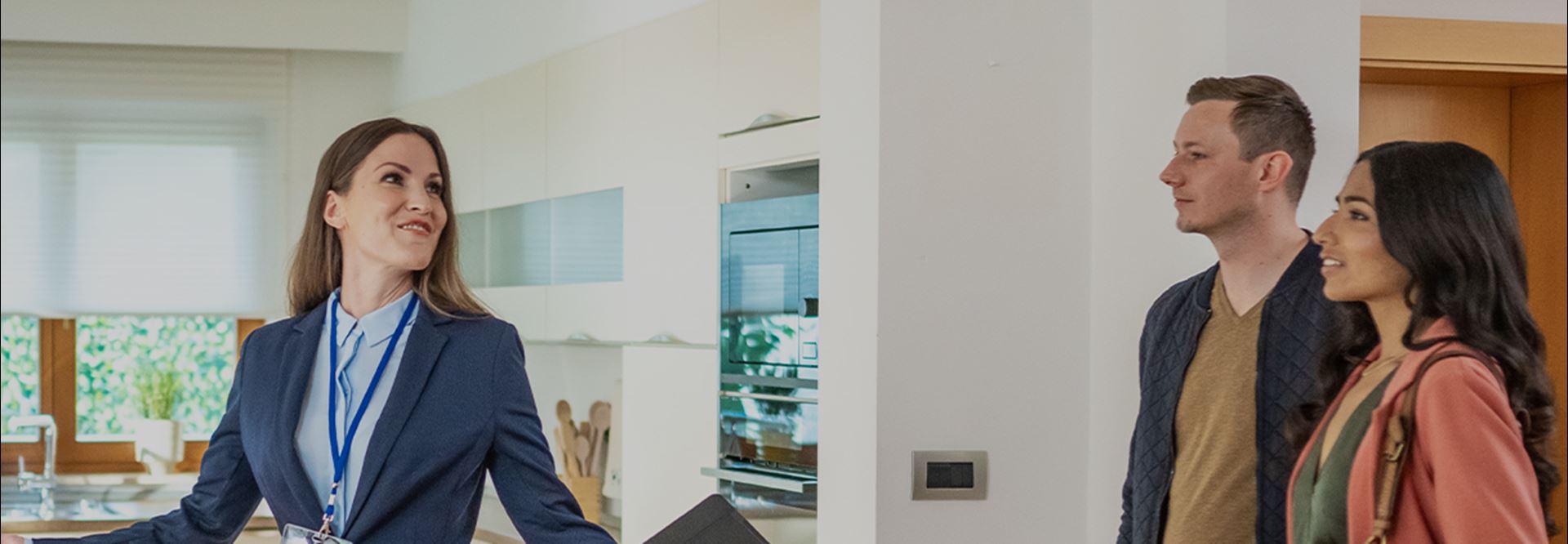 A realtor in a jacket and shirt leads a young couple through the kitchen of a home for sale.