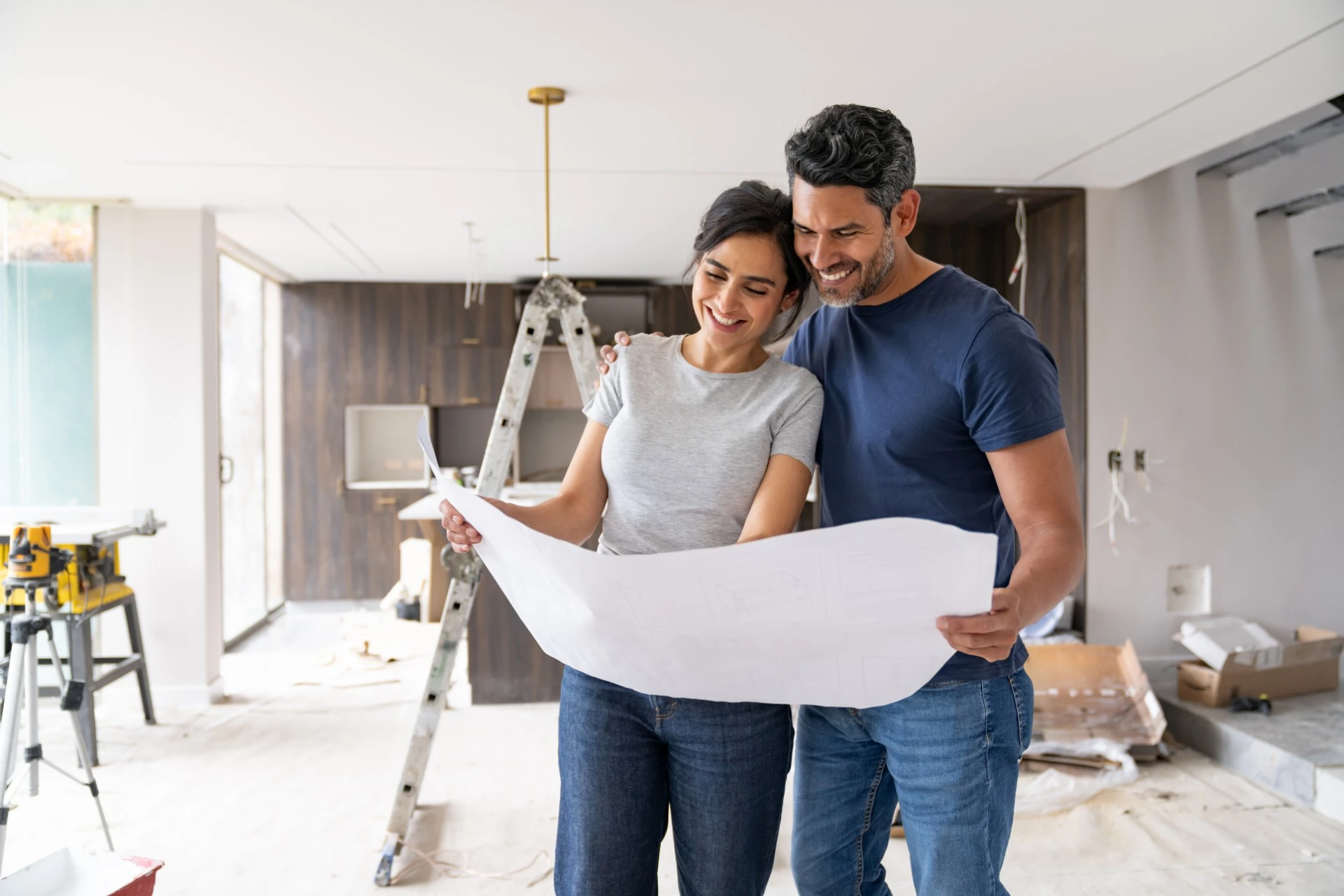 A couple stands together in a room under construction, both hold one end of a large piece of drafting paper.