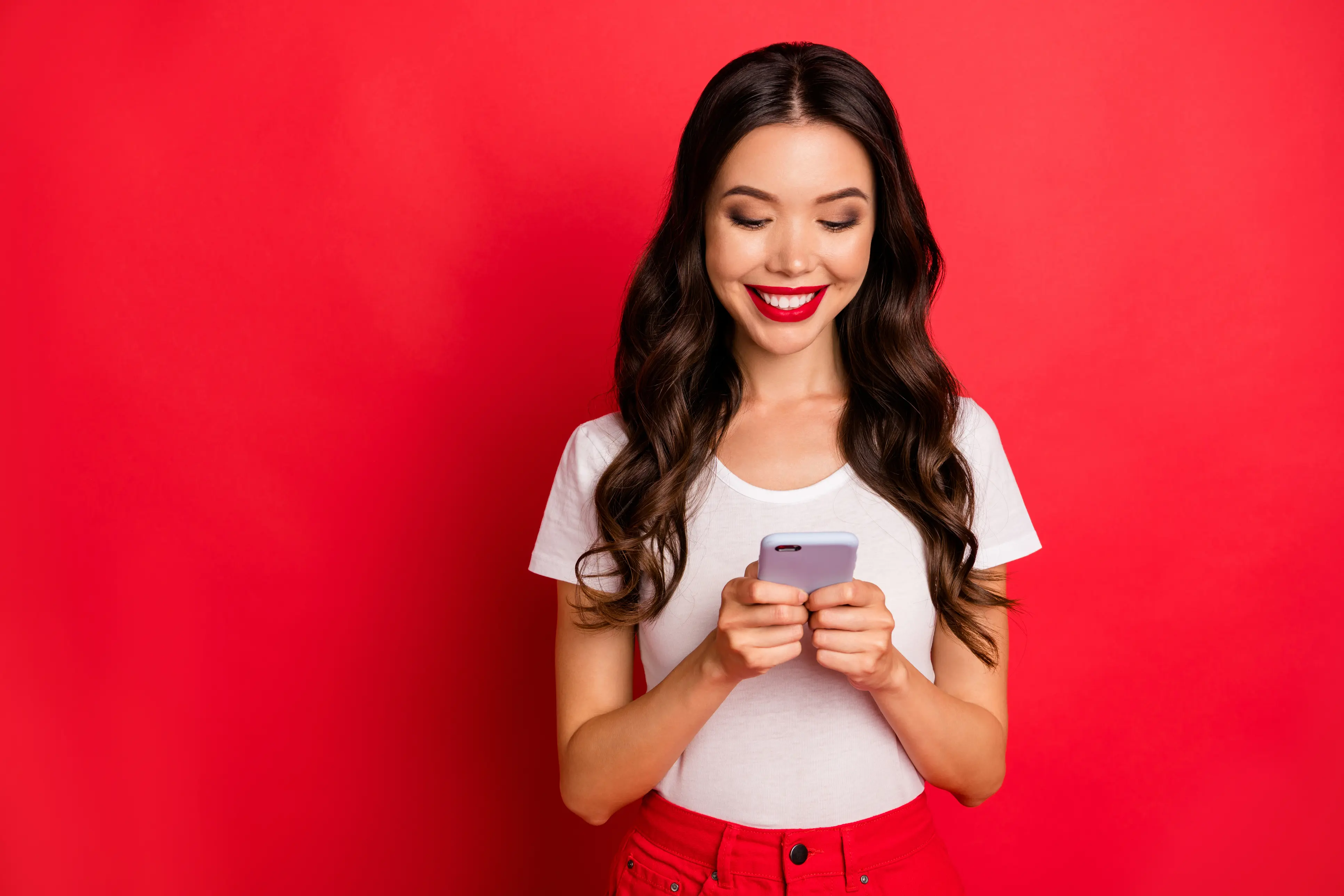 A woman in a white t-shirt stands in front of a red background looking down and smiling at her smartphone that she holds in both hands. A woman in a white t-shirt stands in front of a red background looking down and smiling at her smartphone that she holds in both hands.