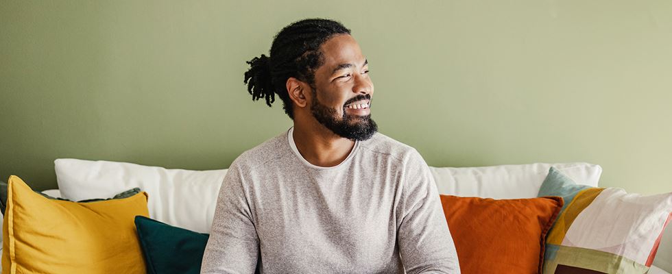A man is pictured sitting on a couch with colorful pillows around him, he looks to his left and smiles.