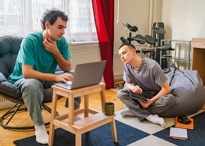Two young men sit next to each other on simple chairs and look intently at a laptop.