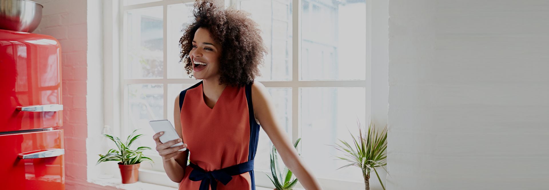 A woman in an orange dress stands in her kitchen and smiles while holding a smartphone in her hand. 