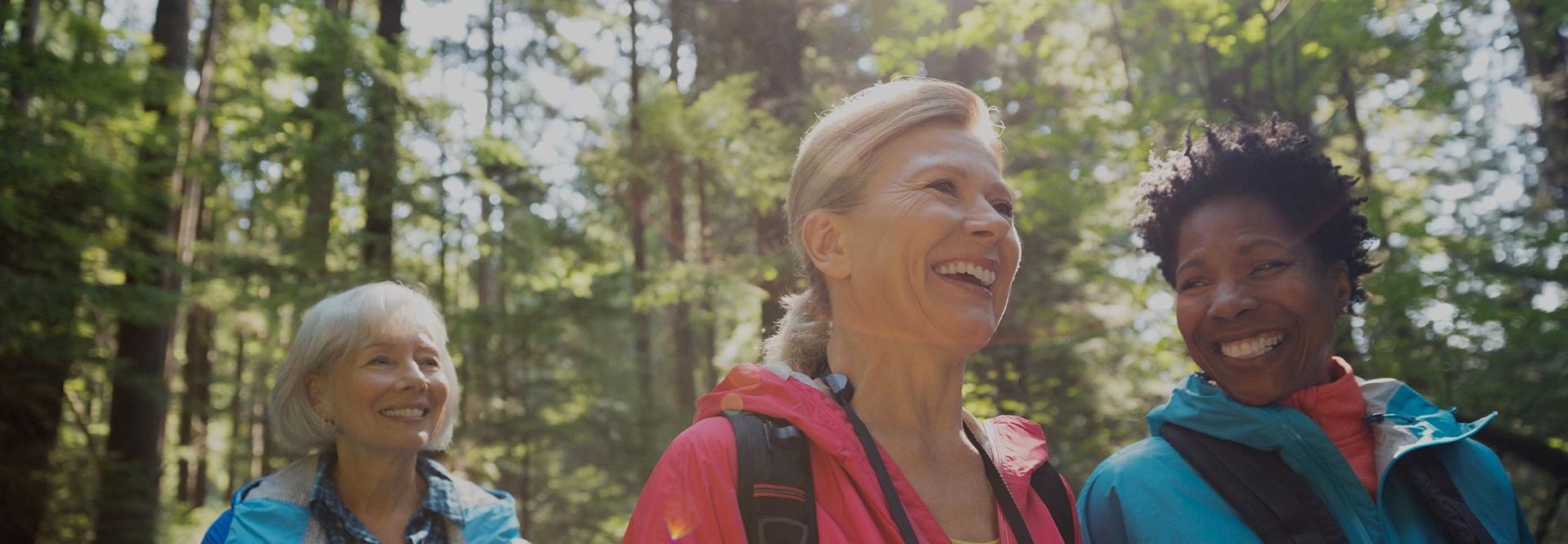 Three smiling elderly woman are hiking in the woods with raincoats and backpacks on.