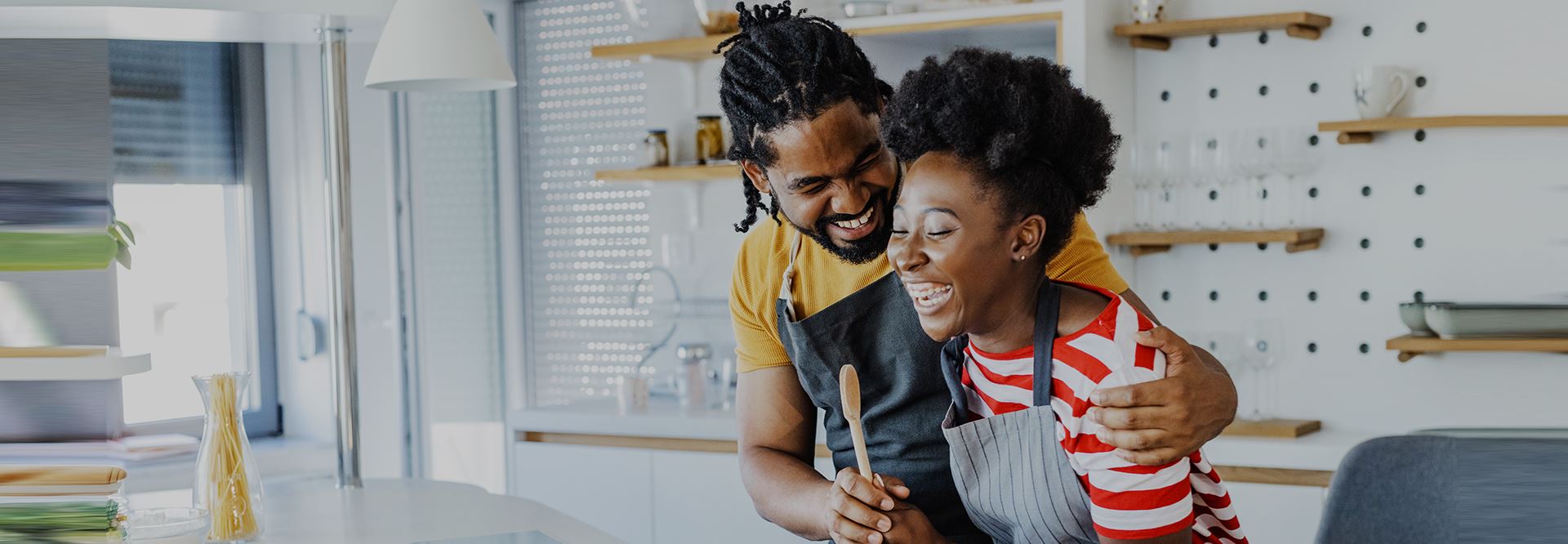 A couple wearing aprons and holding a spoon smile and embrace in their kitchen.