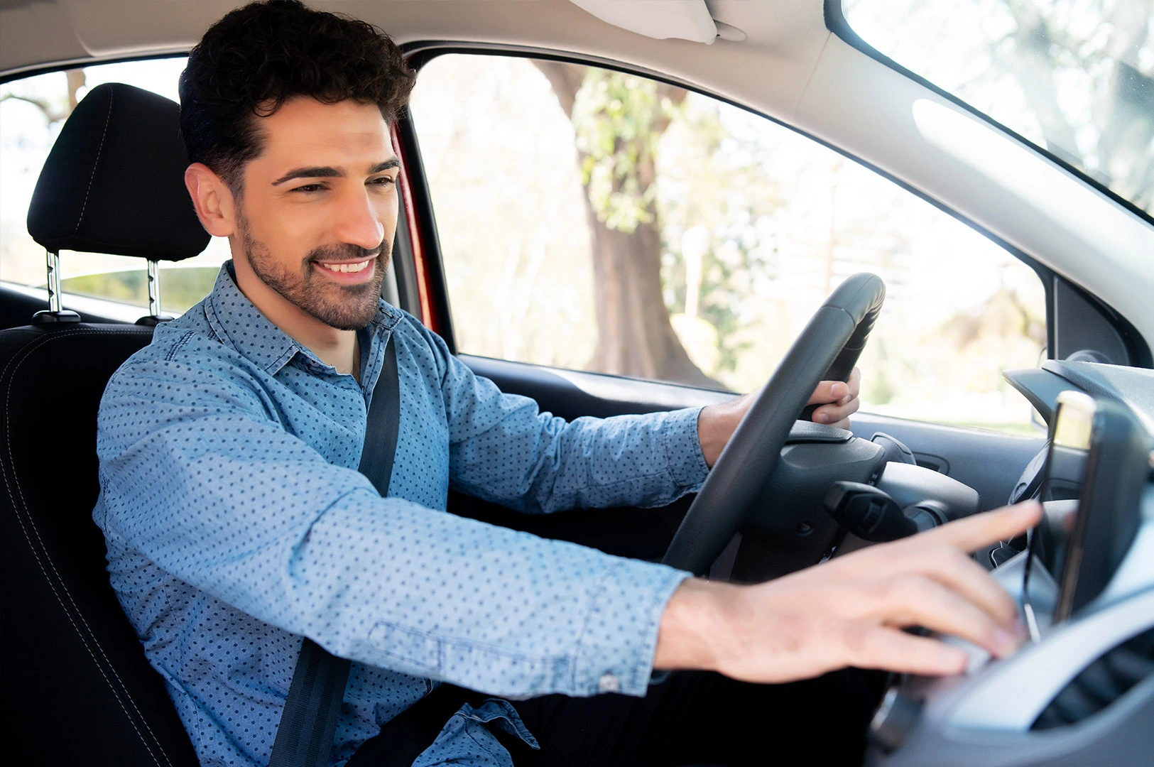 A smiling man is pictured sitting in the driver's seat of his car adjusting the dashboard controls. 