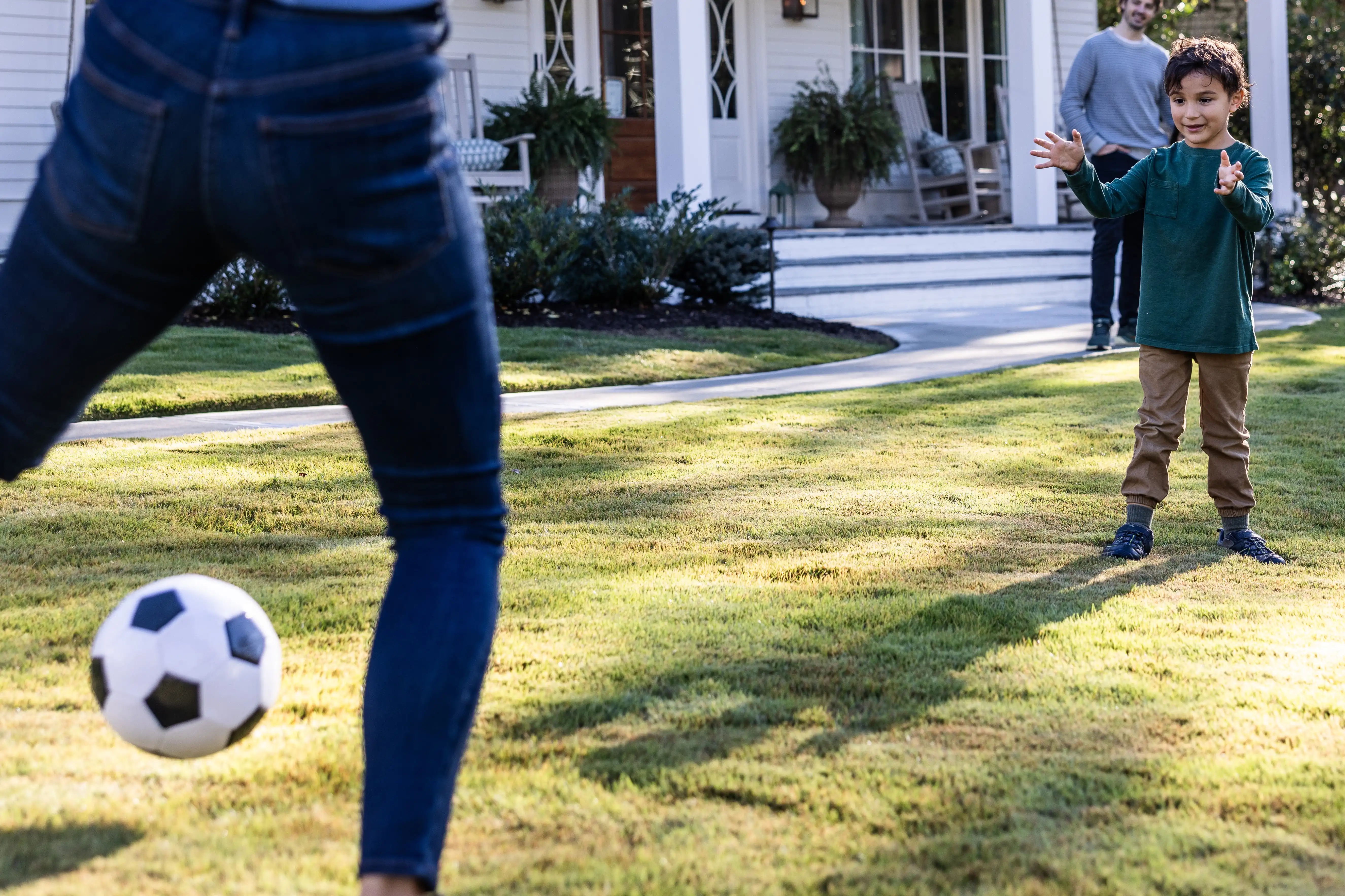 A parent and child kick a soccer ball back and forth in the front yard of their home. 